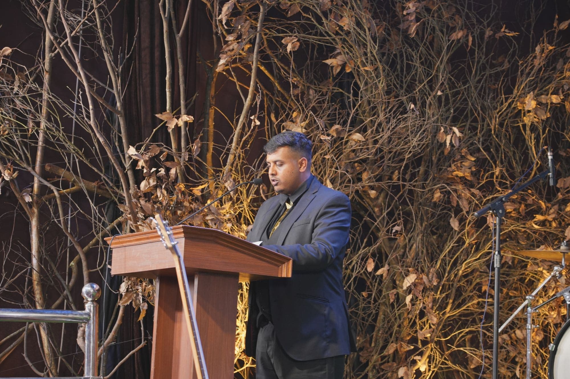 A speaker at the podium on a stage dressed with bare branches and dried leaves